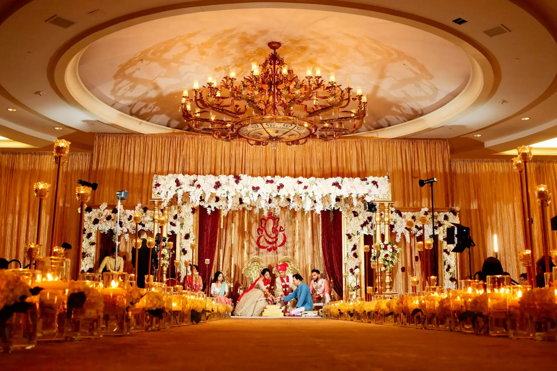 A candle-lit wedding mandap with white flowers, gold pillars and a chandelier