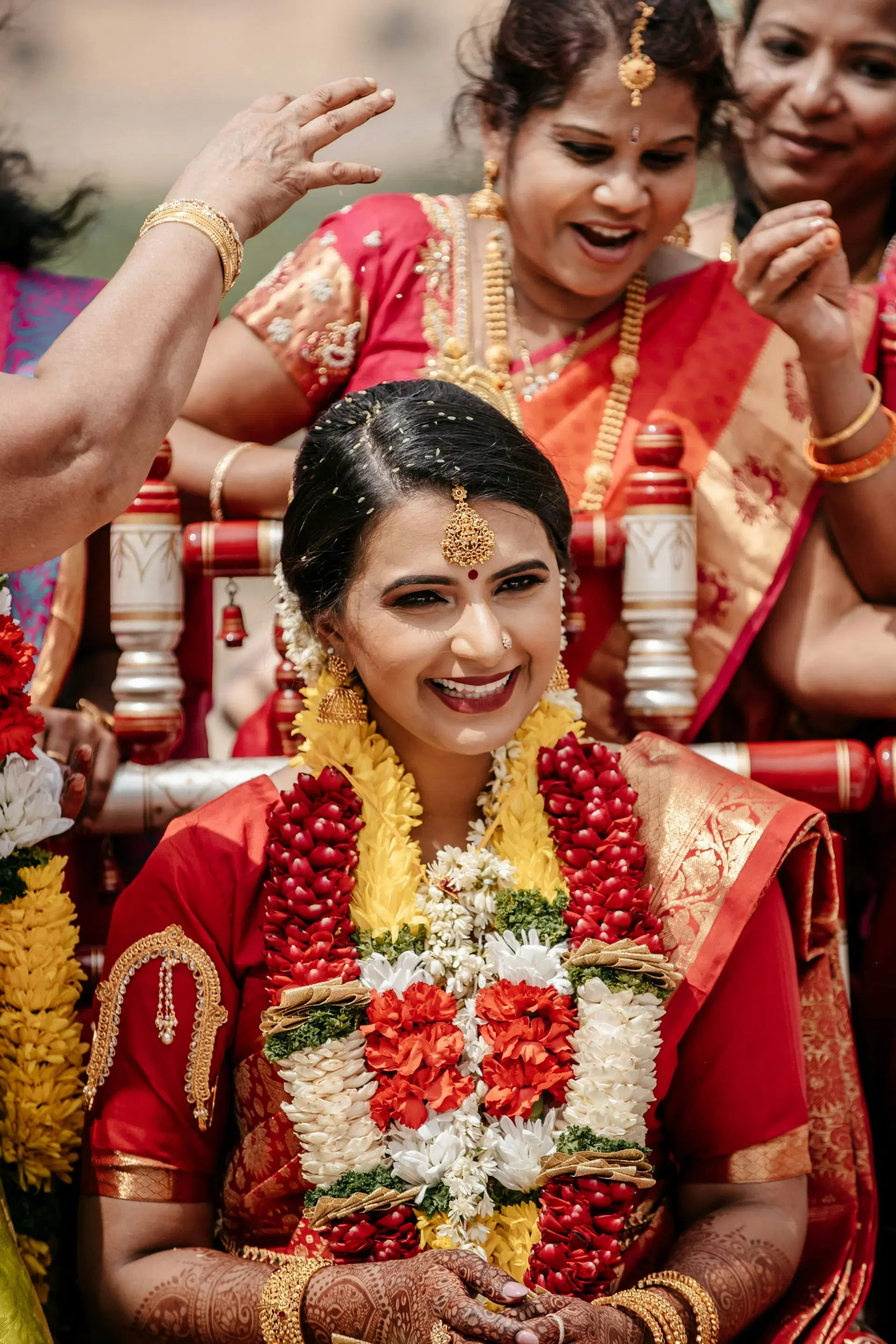 A laughing bride in a red silk saree and flower garlands, with the women of her family blessing her